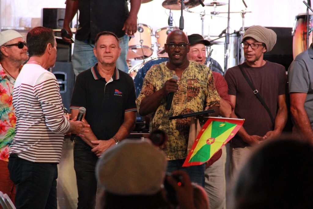 Un représentant de Carriacou et Grenade s’adresse au public, drapeau grenadien à la main, sur la scène du festival
