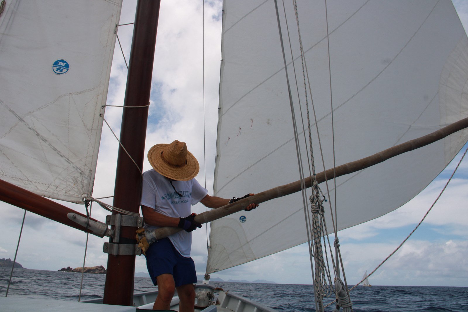 Un équipier manœuvre le tangon en bois contre le mât acajou de Summer Cloud pour tenir le génois ouvert au vent arrière, îlets de Saint-Barthélemy à l’horizon
