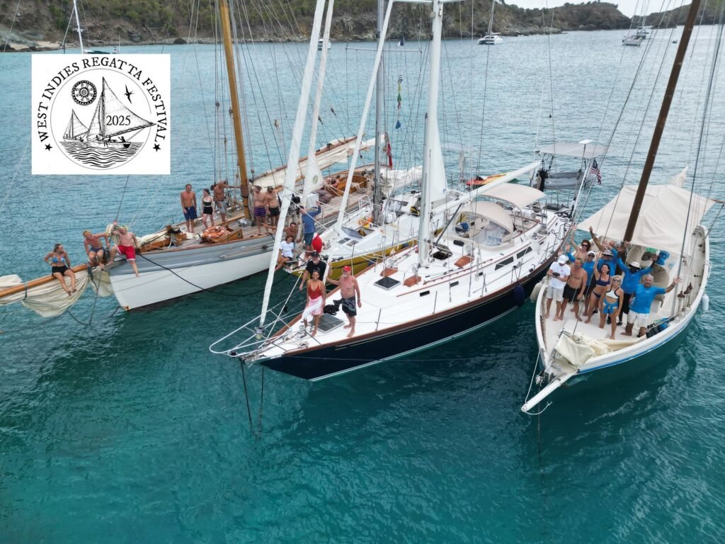 Vue aérienne de quatre voiliers amarrés bord à bord au mouillage pendant la West Indies Regatta Festival 2025, équipages réunis sur les ponts et bras levés, côte de Saint-Barthélemy en arrière-plan.