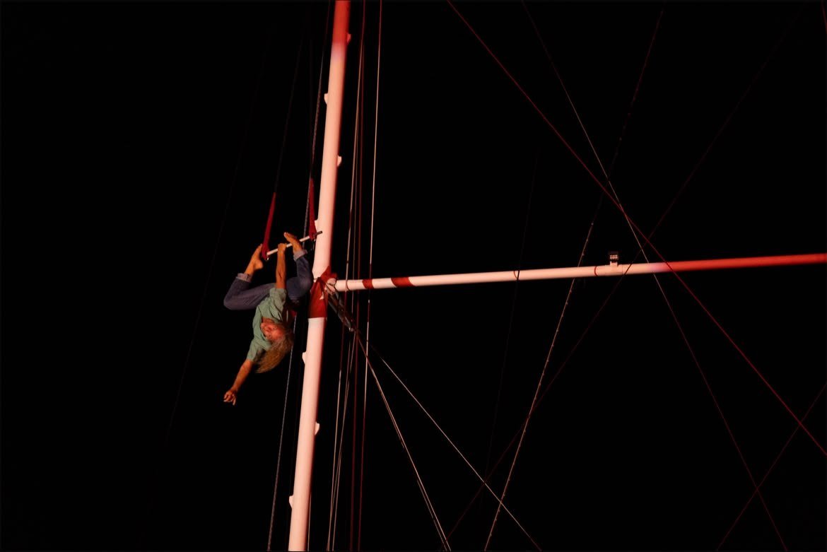 Acrobate suspendu tête en bas à la vergue d'un grand voilier pendant un spectacle nocturne de la West Indies Regatta, mât et gréement éclairés en rouge sur ciel noir.