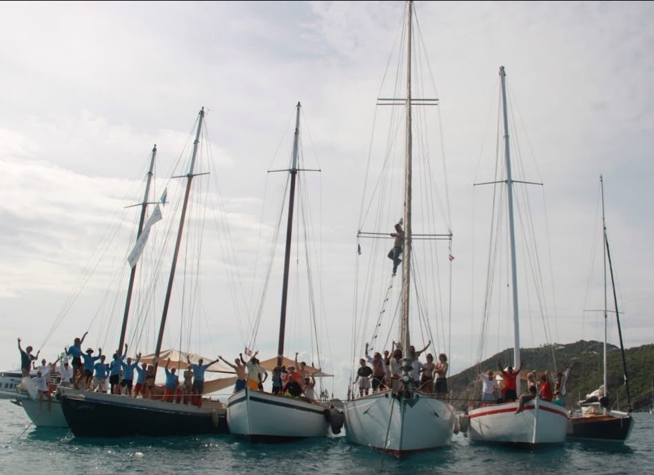 Six sloops traditionnels des Caraïbes rassemblés bord à bord au mouillage pendant la West Indies Regatta, équipages debout sur les ponts bras levés, un marin grimpé dans le mât.