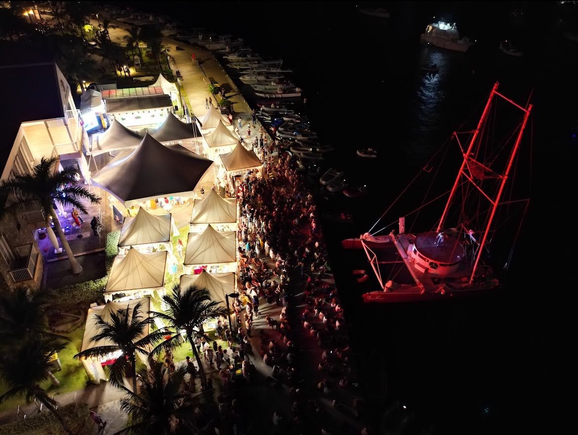 Vue aérienne nocturne du quai de Gustavia pendant la West Indies Regatta Festival : les tentes blanches du marché artisanal, la foule le long du quai et un grand voilier illuminé en rouge à quai.