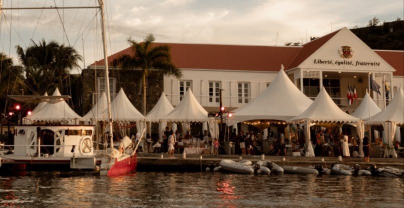 Les tentes blanches du village de la West Indies Regatta alignées sur le quai de Gustavia au crépuscule, devant la mairie de Saint-Barthélemy portant la devise Liberté, égalité, fraternité, un sloop à quai au premier plan.