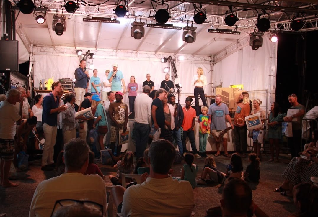 Remise des prix nocturne sous chapiteau : les équipages venus des différentes îles des Caraïbes rassemblés sur scène avec leurs trophées peints à la main, devant un public assis.