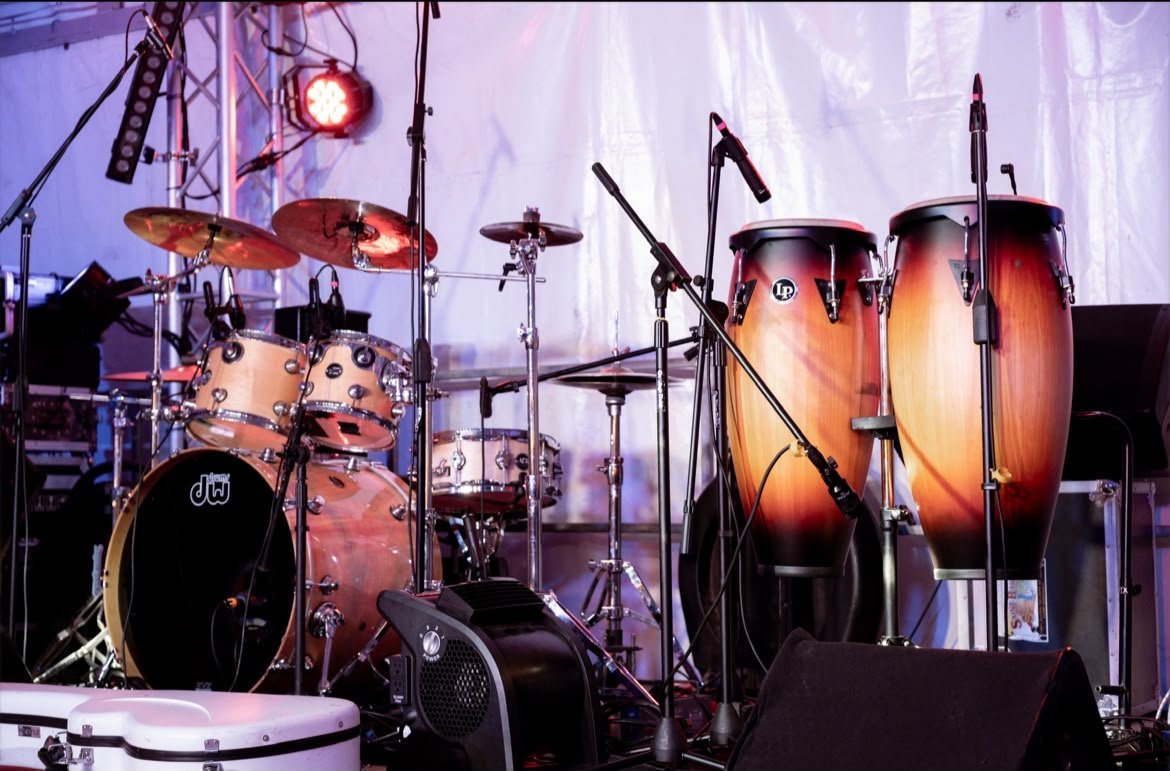 Scène de concert du festival avant le début du set : batterie, congas et cymbales sous les projecteurs, micros et retours installés devant la toile blanche du chapiteau.