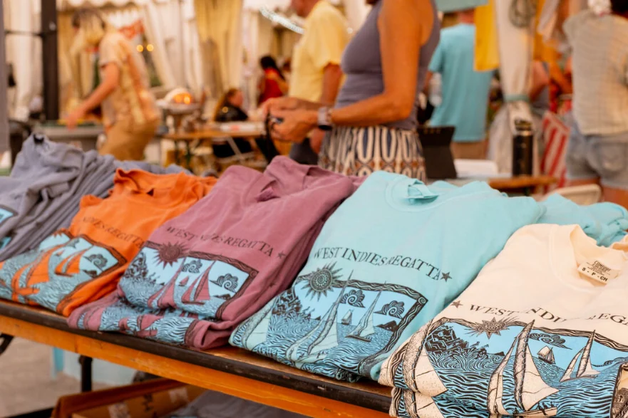 Tee-shirts de la West Indies Regatta 2026 pliés sur une table du marché, imprimés du dessin d’Aragorn Dick-Read