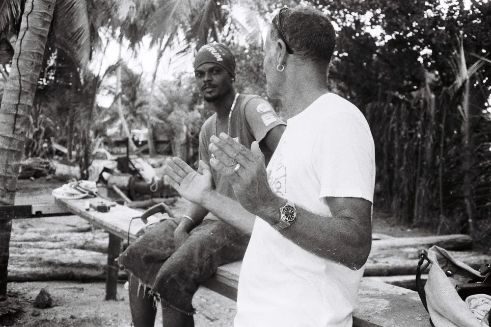 Alexis Andrews discute avec Callistus Enoe assis sur un établi du chantier