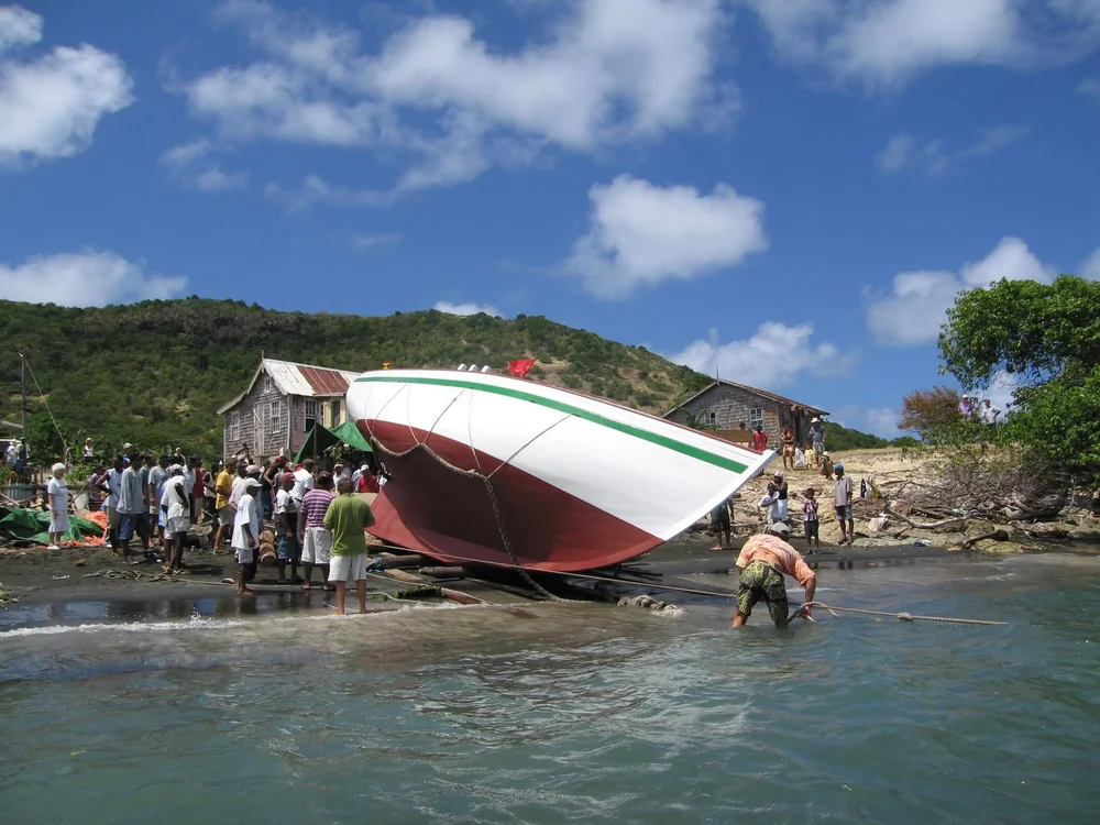 La coque rouge et blanche de Genesis, couchée sur la plage de Windward, entourée du village ; un homme tire une aussière dans l’eau