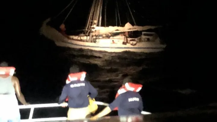 Le sloop Mermaid de nuit au large de La Ceiba, coque blanche éclairée par les projecteurs, gîté dans les brisants ; au premier plan, des sauveteurs en gilet de sauvetage à bord d’un canot