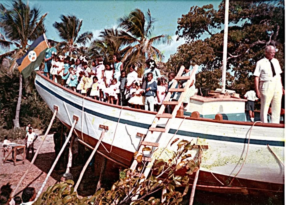 Les enfants du village, en habits du dimanche, alignés à bord du sloop Mermaid encore posé à terre le jour de son lancement, une échelle appuyée au bordé et un pavois hissé à l’avant