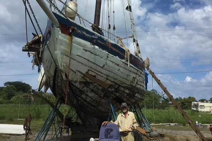 Le sloop Mermaid of Carriacou au sec sur ses bers dans un chantier, la coque en bois décapée, son capitaine debout au pied de la quille