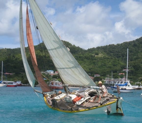 Le cotre Plumbelly sous voiles, grand-voile aurique et deux focs, barré par un homme au chapeau de paille dans un mouillage des Antilles